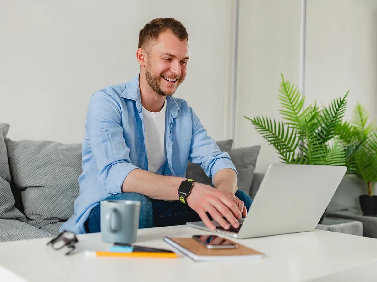 handsome-smiling-man-sitting-sofa-drinking-tea-home-table-working-online-laptop-from-home